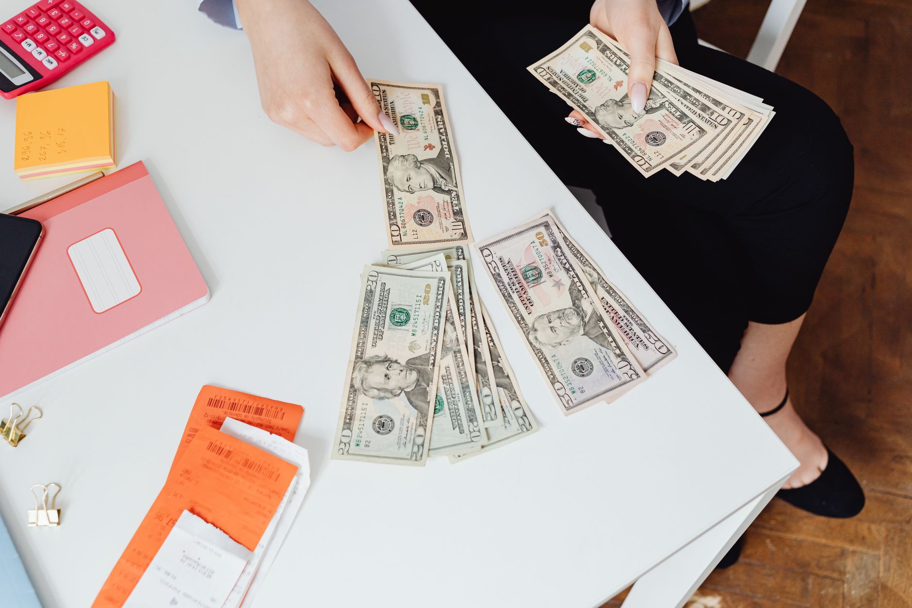 A person counting US dollar bills at a white desk with office supplies nearby.
