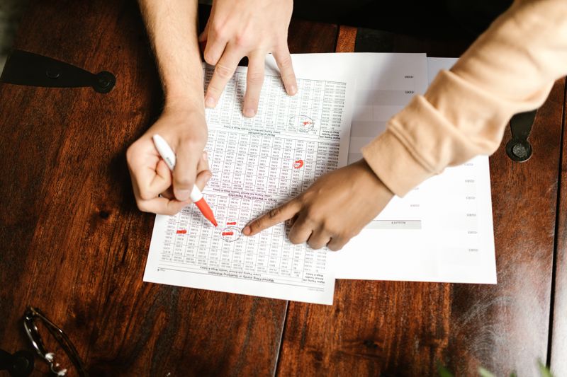Quanto custa manter uma estrutura offshore por ano Two hands reviewing and marking financial documents with a red pen on a wooden table, top view.