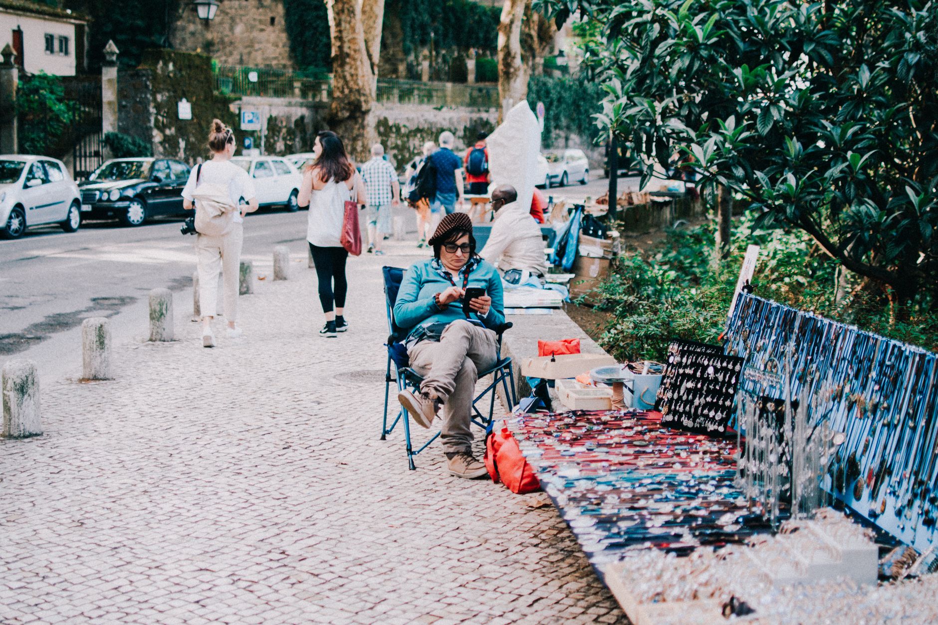 A woman selling handmade jewelry on a sidewalk in a lively urban park, engaging with tourists.