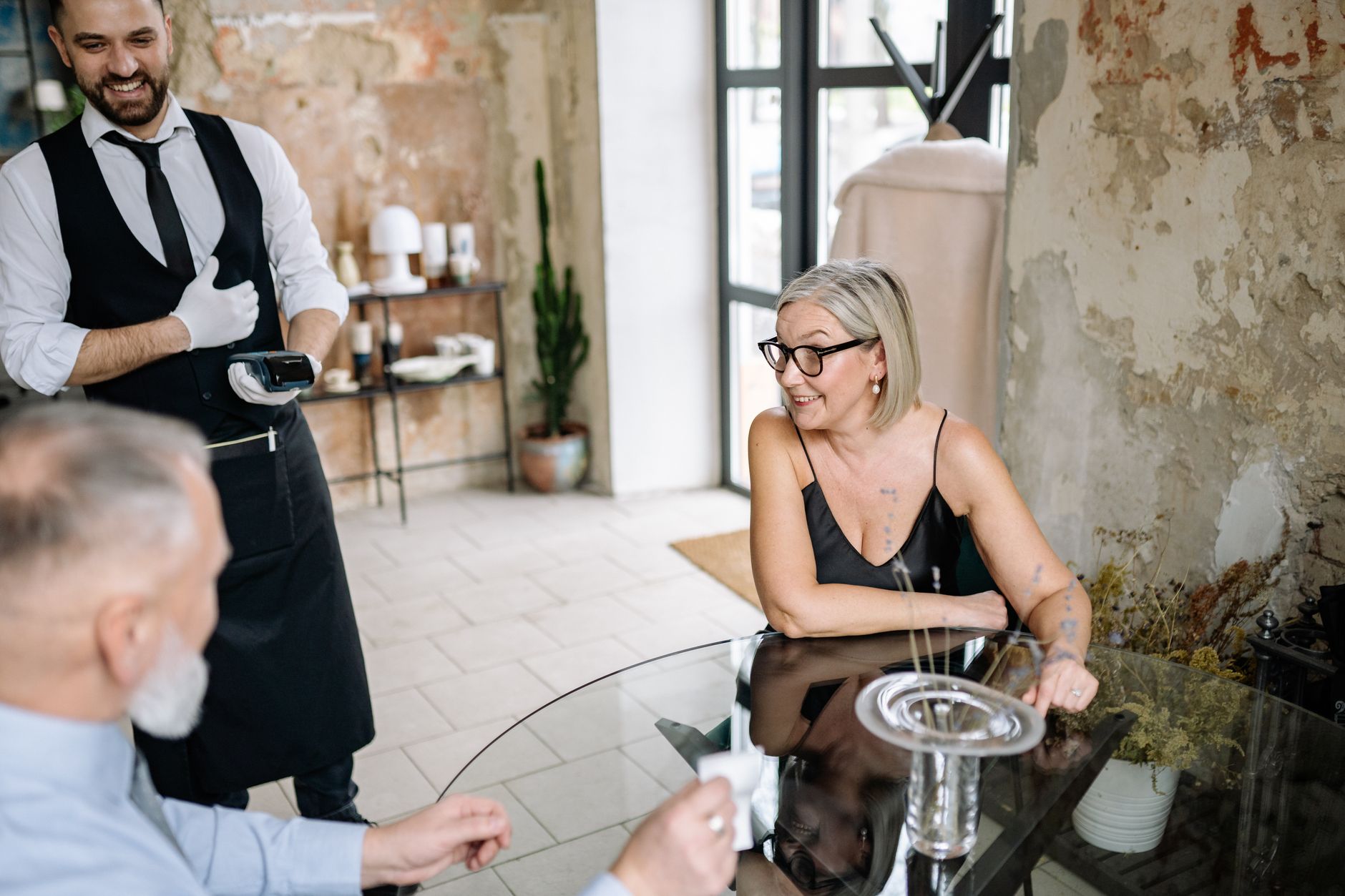Quantos euros levar para uma viagem? A waiter serves a senior couple in a rustic café, highlighting elegance and enjoyment.