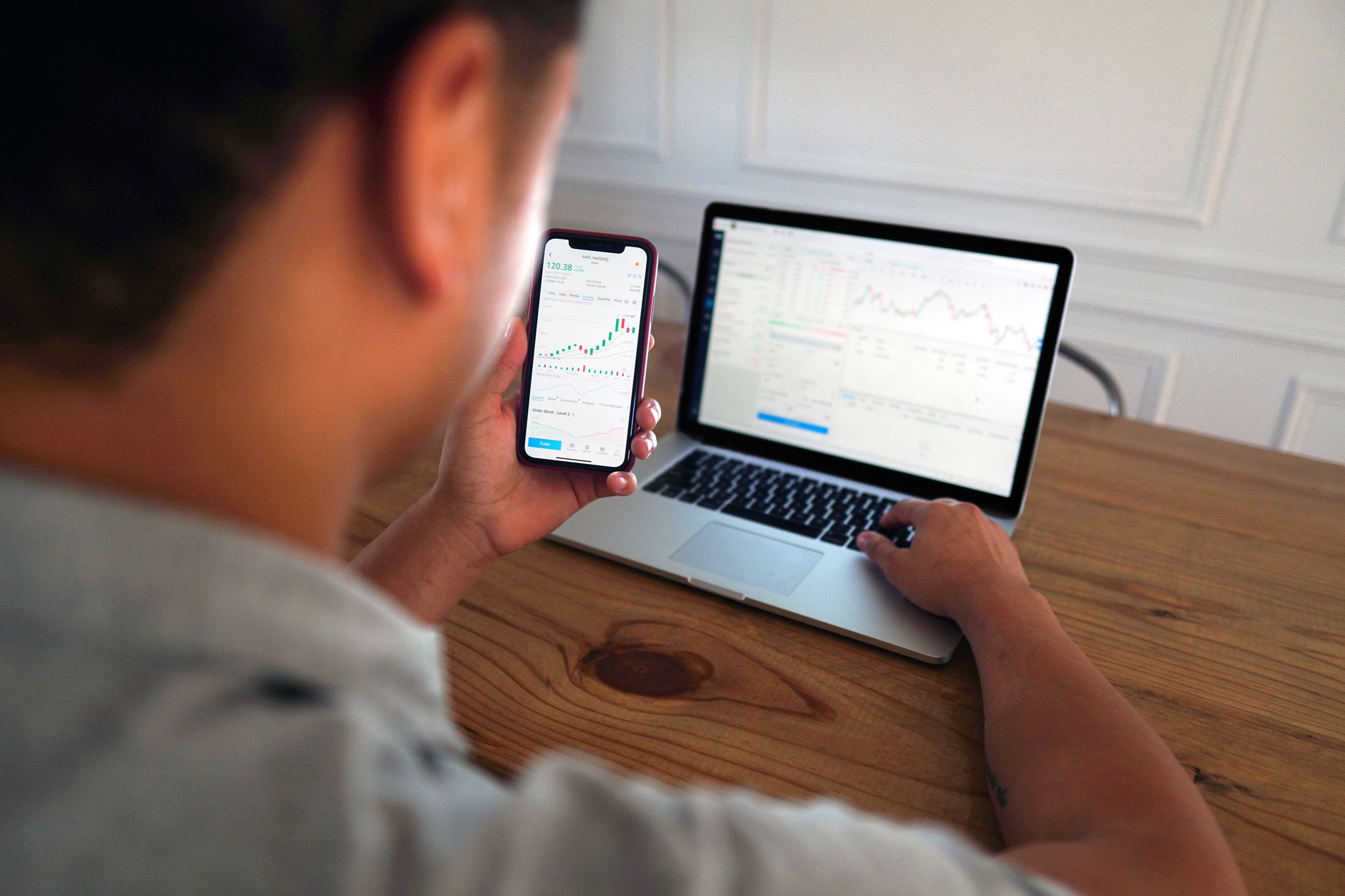 A man sits at a wooden table analyzing stock market trends using a laptop and smartphone.