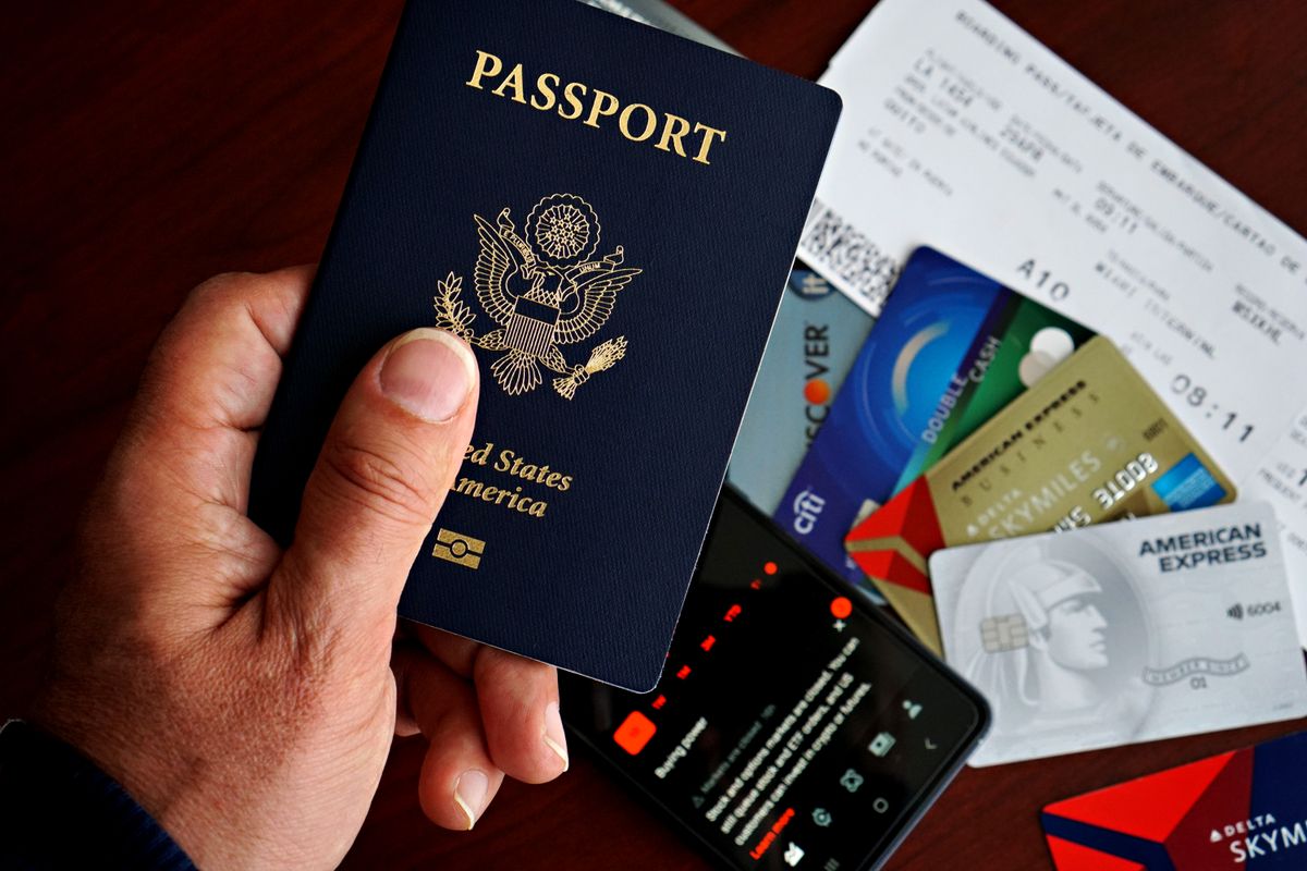 Pessoa segurando um passaporte e um cartão de débito internacional diante de um notebook em uma mesa.