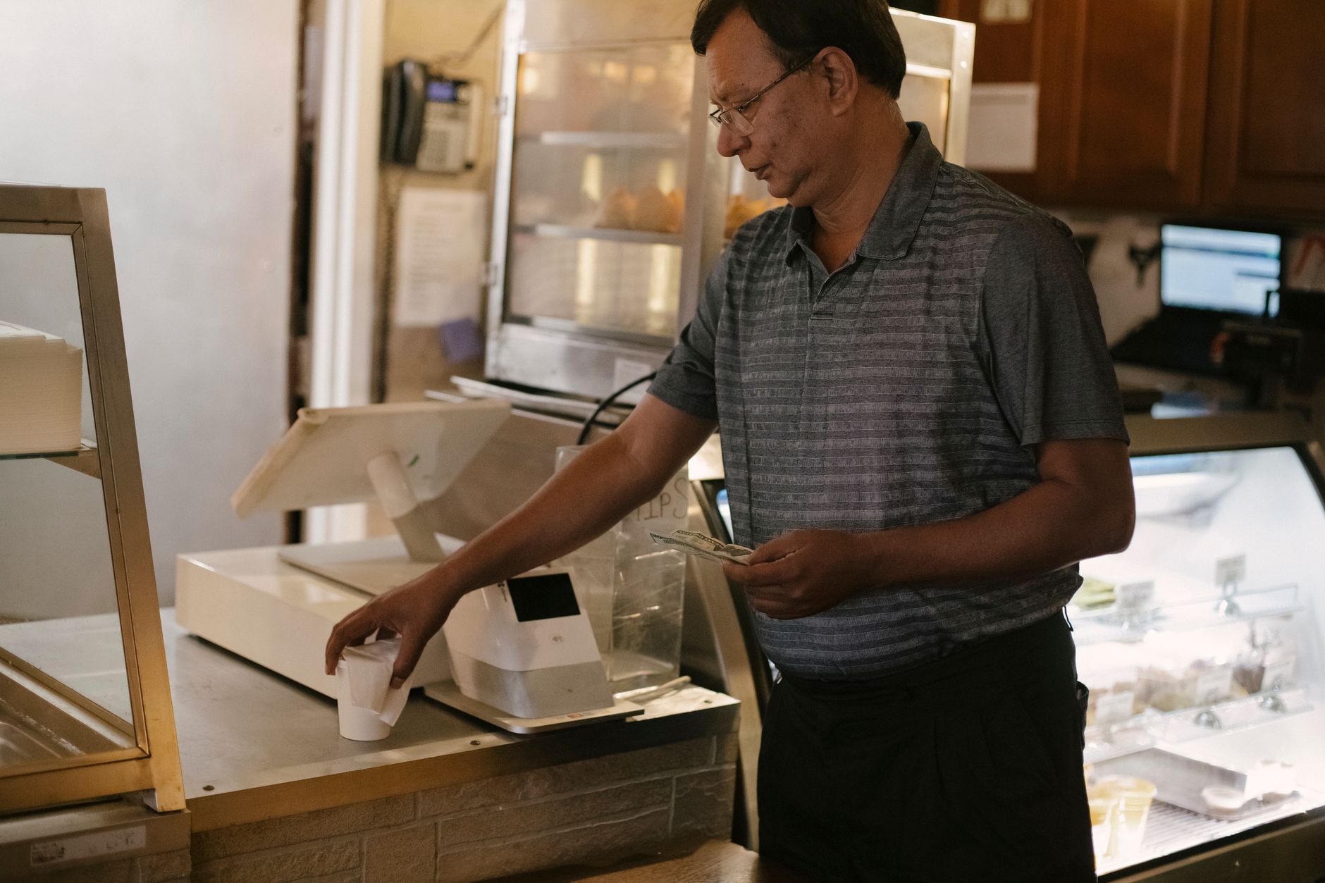 Cupom fiscal e nota fiscal são a mesma coisa? Side view of Asian male wearing eyeglasses standing near cash register and showcase while piking up cup of coffee in cafe