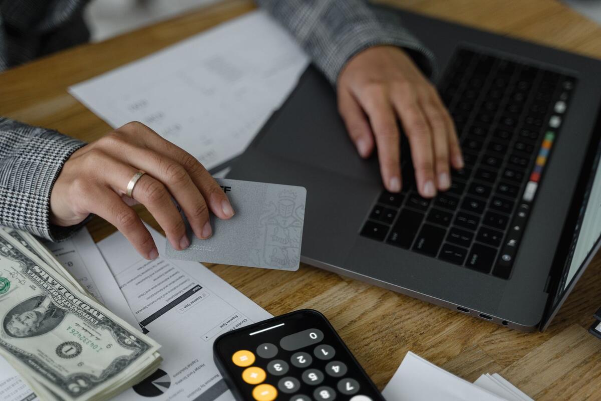 Pessoa usando cartão de crédito e notebook ao lado de calculadora e dinheiro