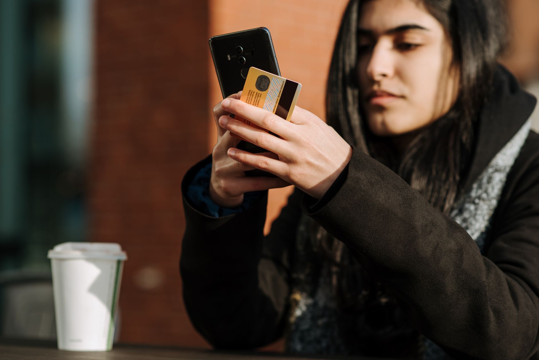 Cartão sem consulta ao SPC existe de verdade? Crop young Hispanic female shopper with debit card making purchase on cellphone near takeaway beverage in cafeteria