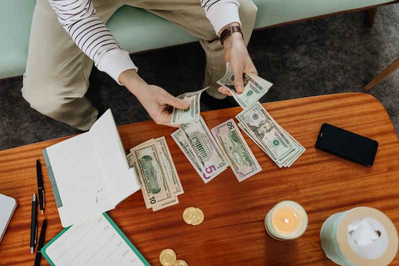 Como criar o hábito de poupar todo mês, mesmo pouco A person sitting and counting various US dollar bills at a table indoors.