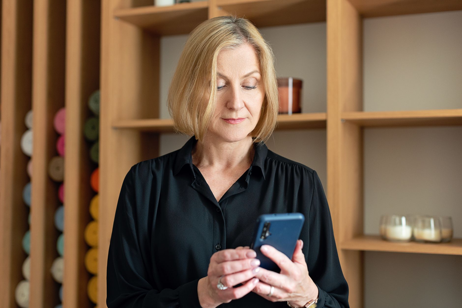 Professional woman in black attire using a smartphone indoors with shelving background.