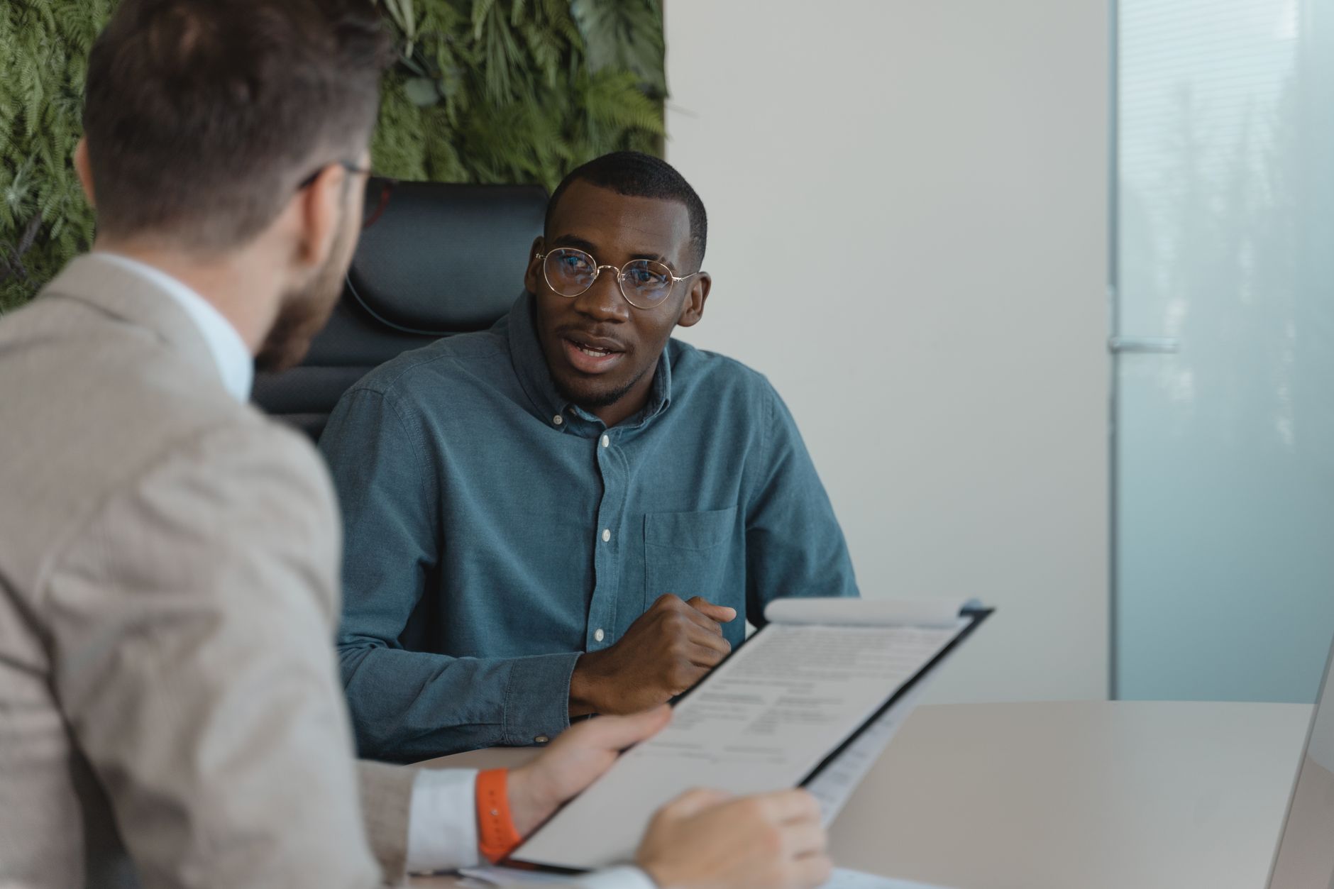 Consignado privado: o que é e quem pode contratar Two men engaged in a professional meeting at an office table with documents and a plant wall background.