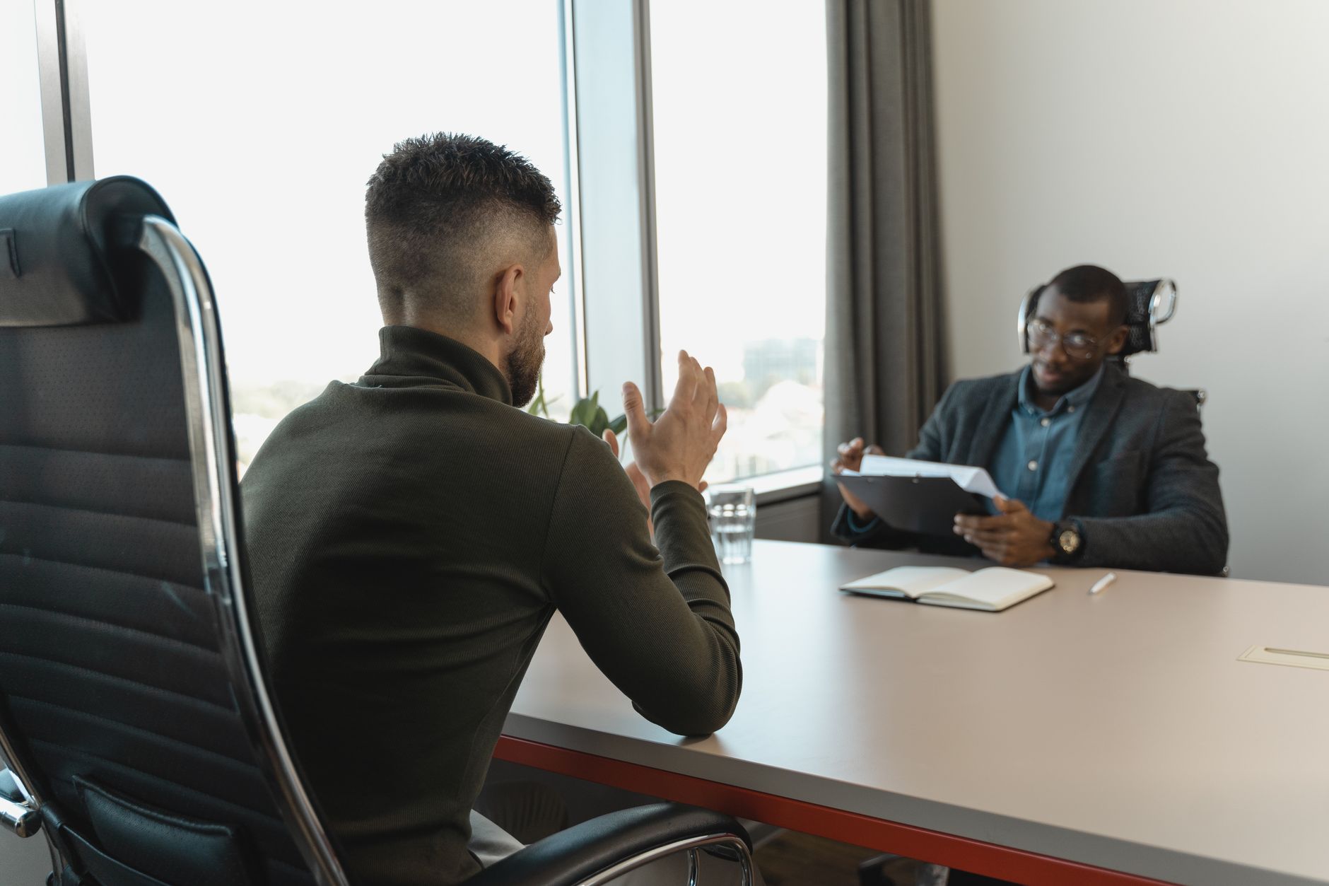 Consignado para CLT: como funciona o desconto em folha Two men in a job interview setting in a modern office with large windows.