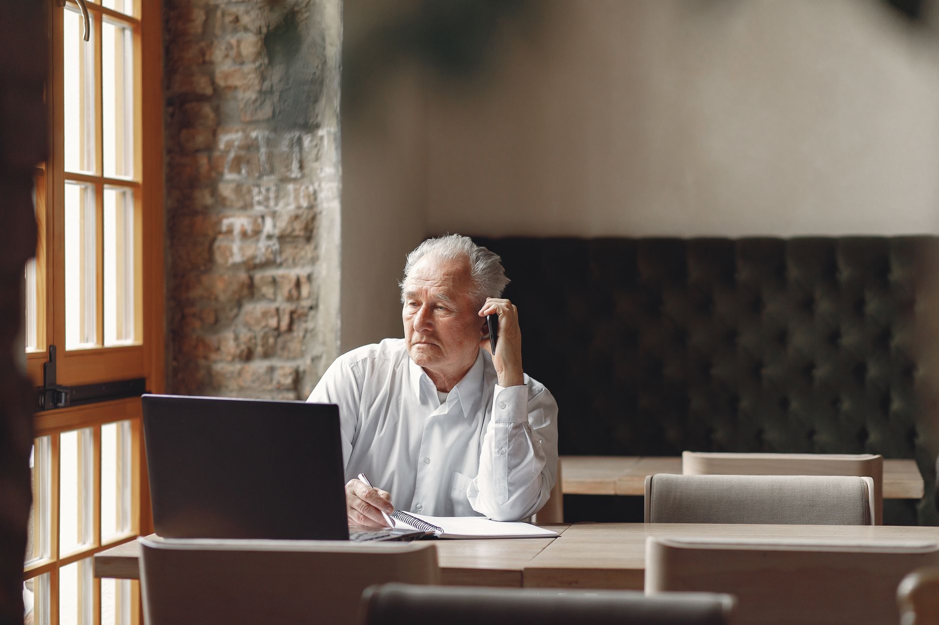 Empréstimo consignado do INSS: como funciona Thoughtfull senior male calling on smartphone and taking notes while working remotely in creative modern workspace at daytime and looking at window