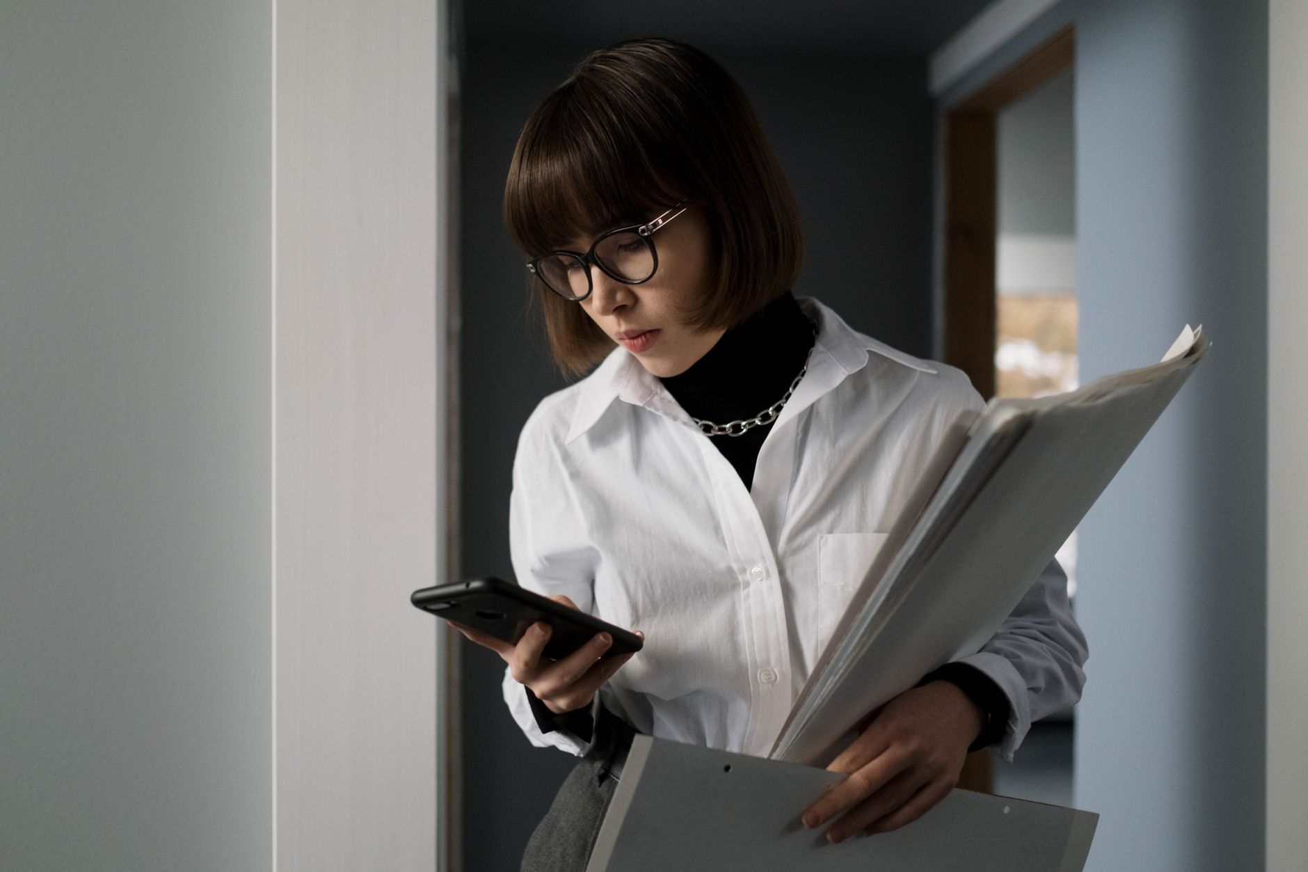 Young woman with eyeglasses, holding documents, checking smartphone indoors.