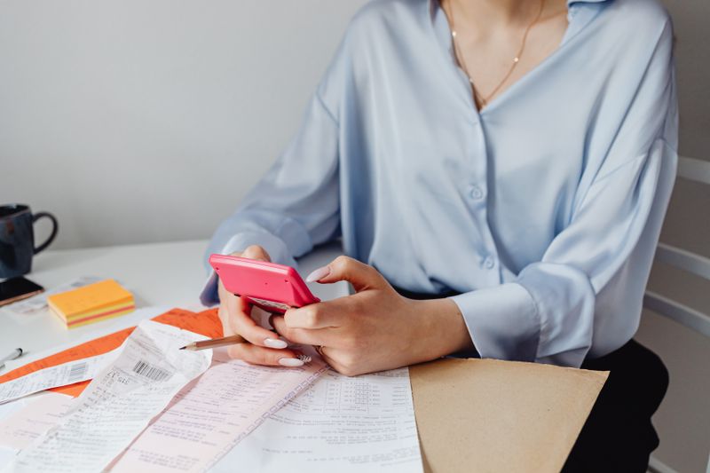 Vale a pena investir antes de quitar as dívidas? A woman using a pink calculator surrounded by bills and receipts at a desk.