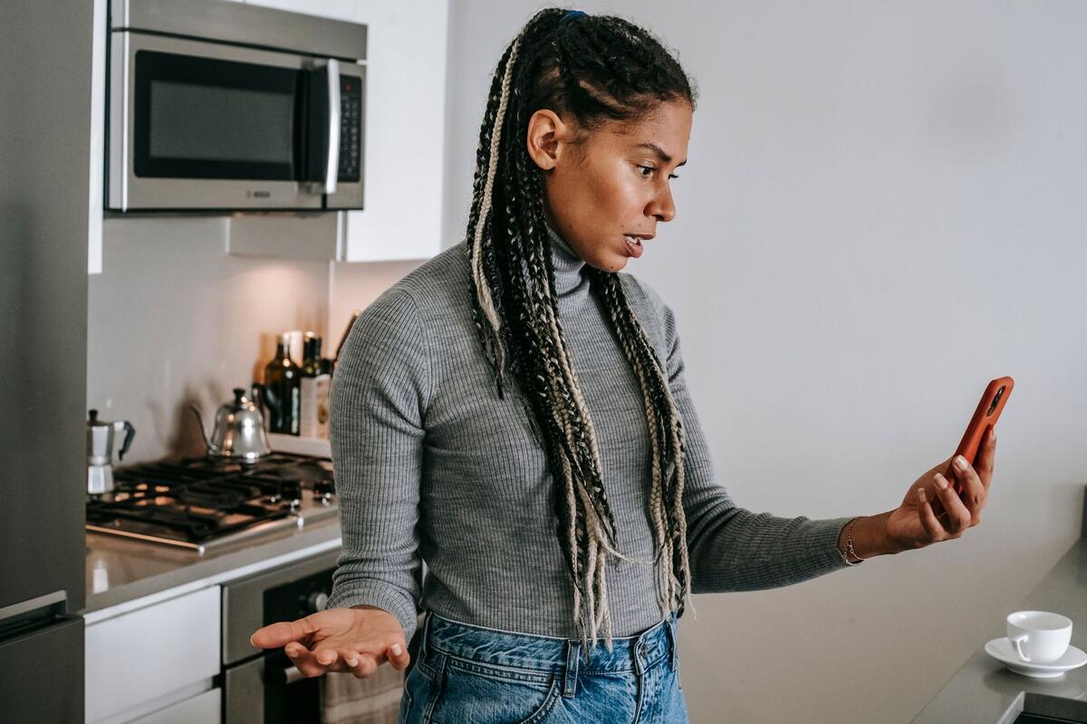 O banco precisa avisar antes? Mulher com expressão séria olhando o celular na cozinha