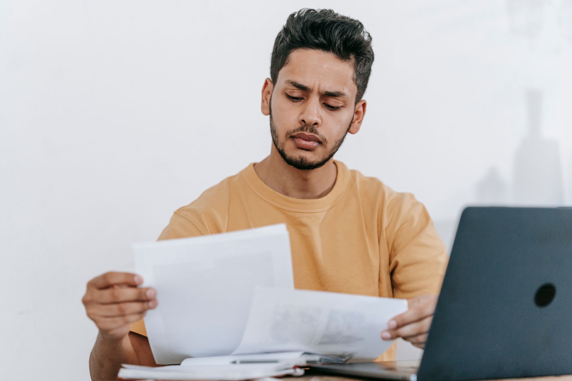 Margem consignável: o que é e como consultar Attentive young bearded Hispanic male remote worker examining papers with results of project while working with laptop in home office