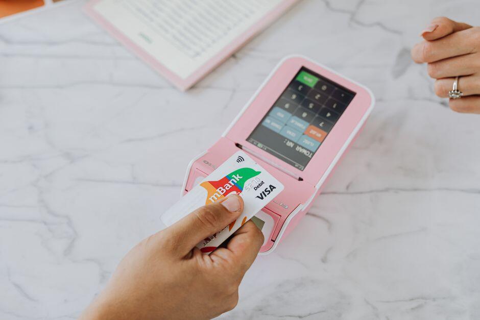 Mão aproximando cartão de uma maquininha de pagamento rosa