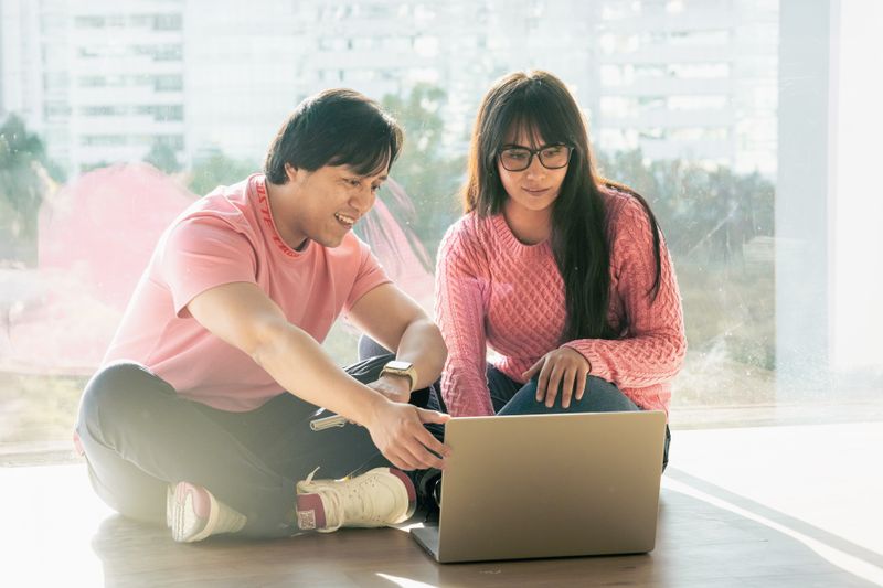Two young adults in casual attire working together on a laptop by a window.