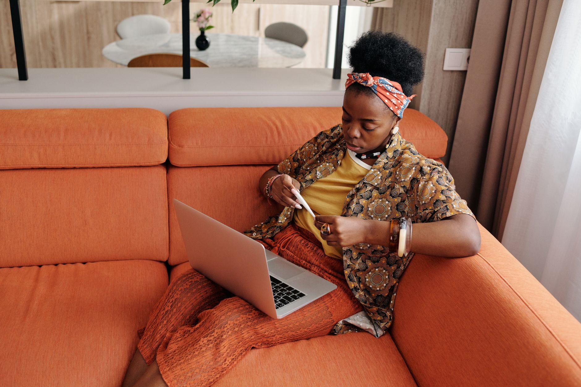 Score baixo: ainda dá para conseguir empréstimo? A worried woman sitting on an orange sofa checking her temperature with a thermometer, while a laptop sits open beside her.