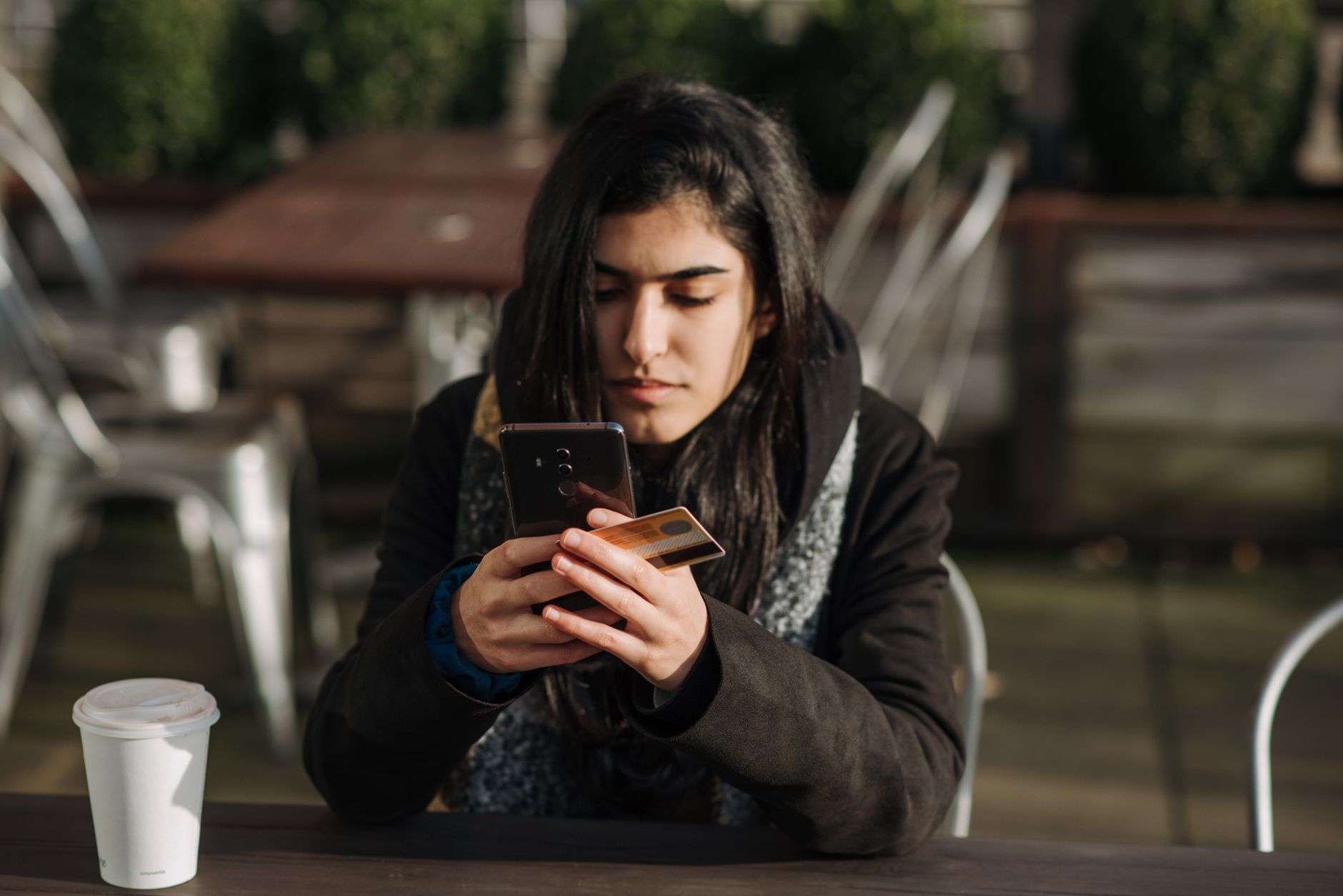 Serasa Limpa Nome: como funciona e como usar Young ethnic female purchaser with credit card shopping online on mobile phone at table with takeaway hot drink in cafeteria