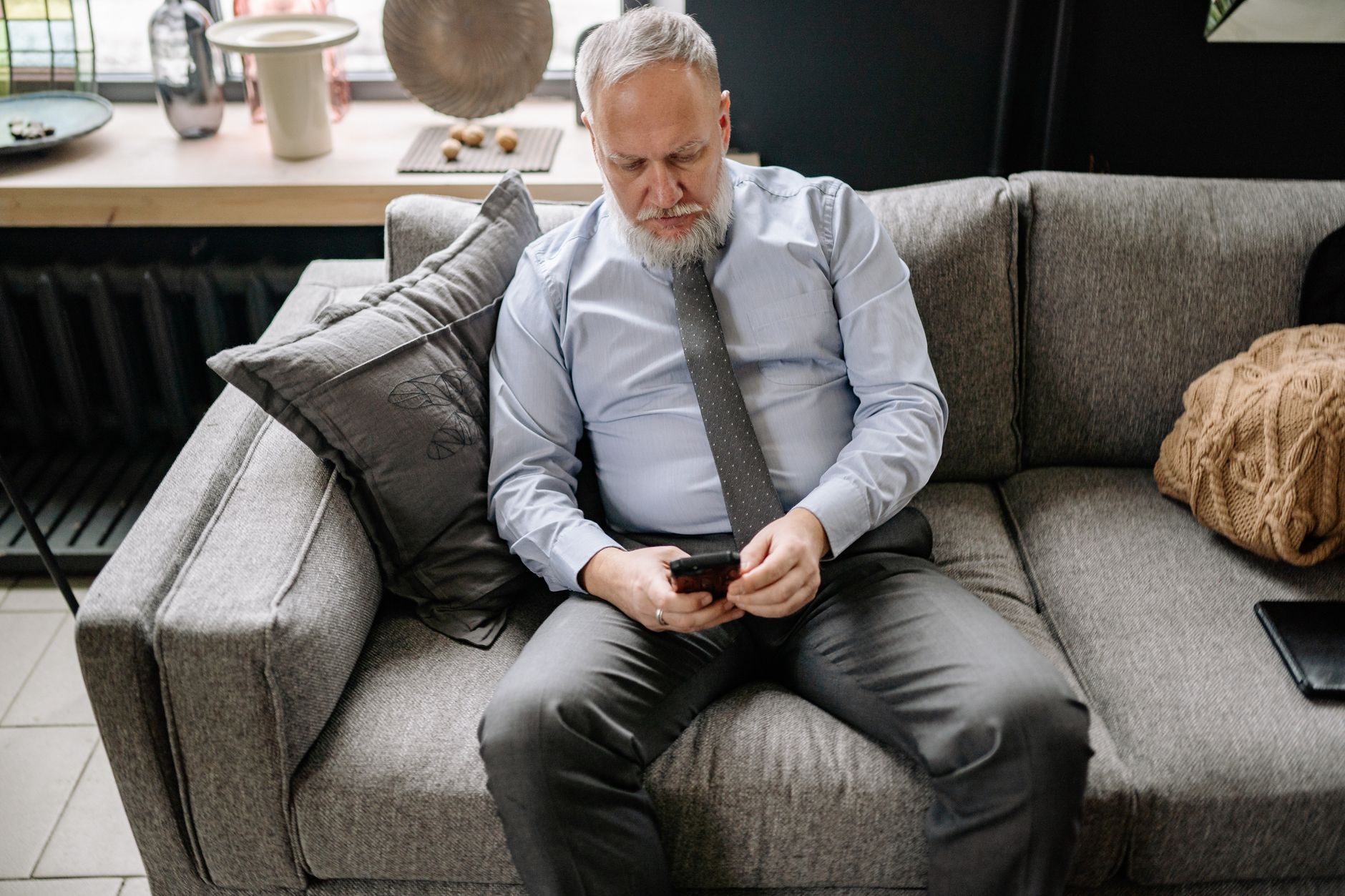 Troco de consignado: o que significa na portabilidade Senior man in formal attire scrolling on phone while sitting on a couch.