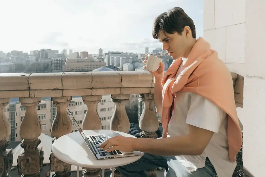 Homem tomando café e trabalhando em um notebook em uma varanda com vista da cidade Homem tomando café e trabalhando em um notebook em uma varanda com vista da cidade