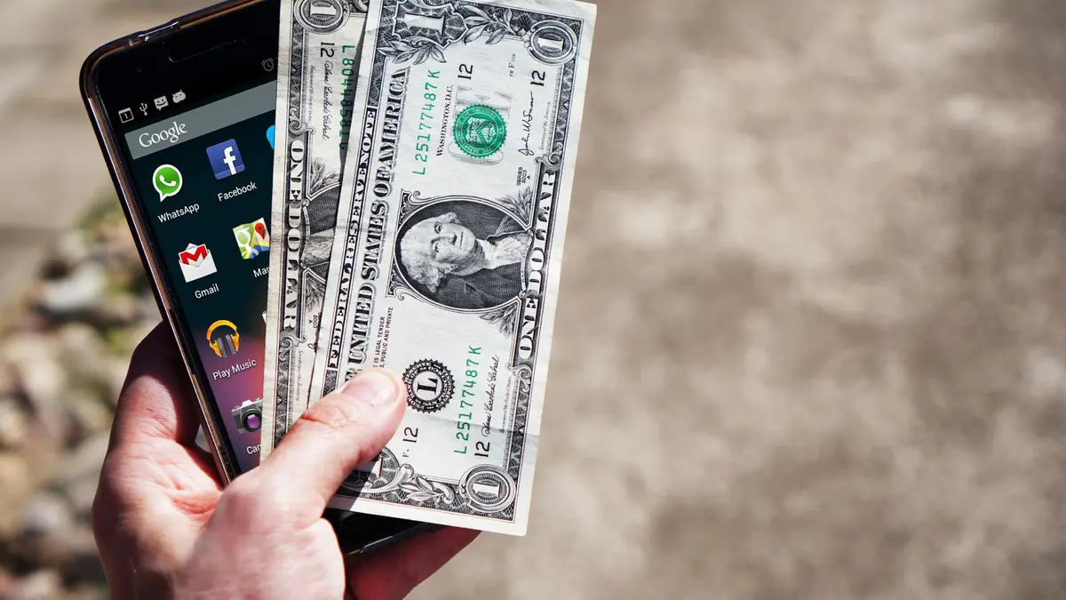 Mão segurando celular e notas de dólar