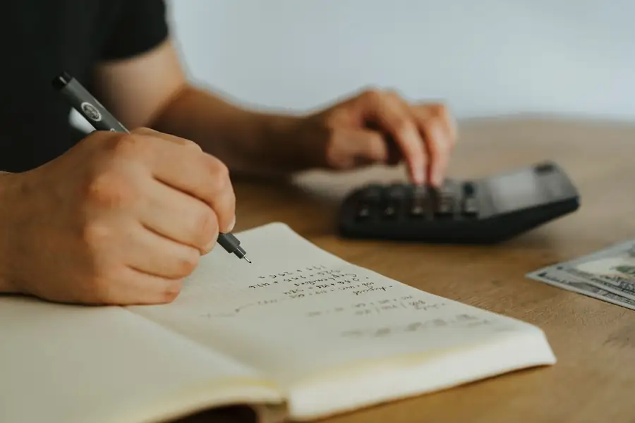 Mãos fazendo cálculos em um caderno ao lado de uma calculadora
