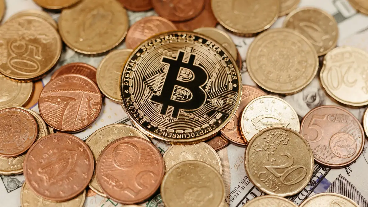 Moedas digitais e notas de dólar e euro sobre mesa com notebook