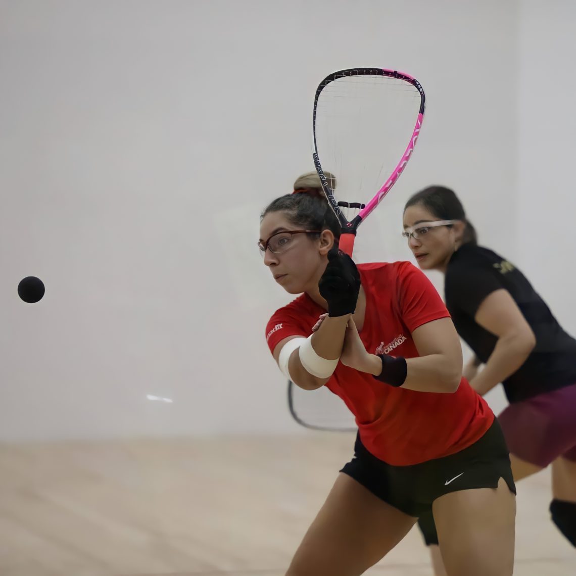 Michèle Morissette is hitting a racquetball with a backhand swing. Her opponent is in the background.