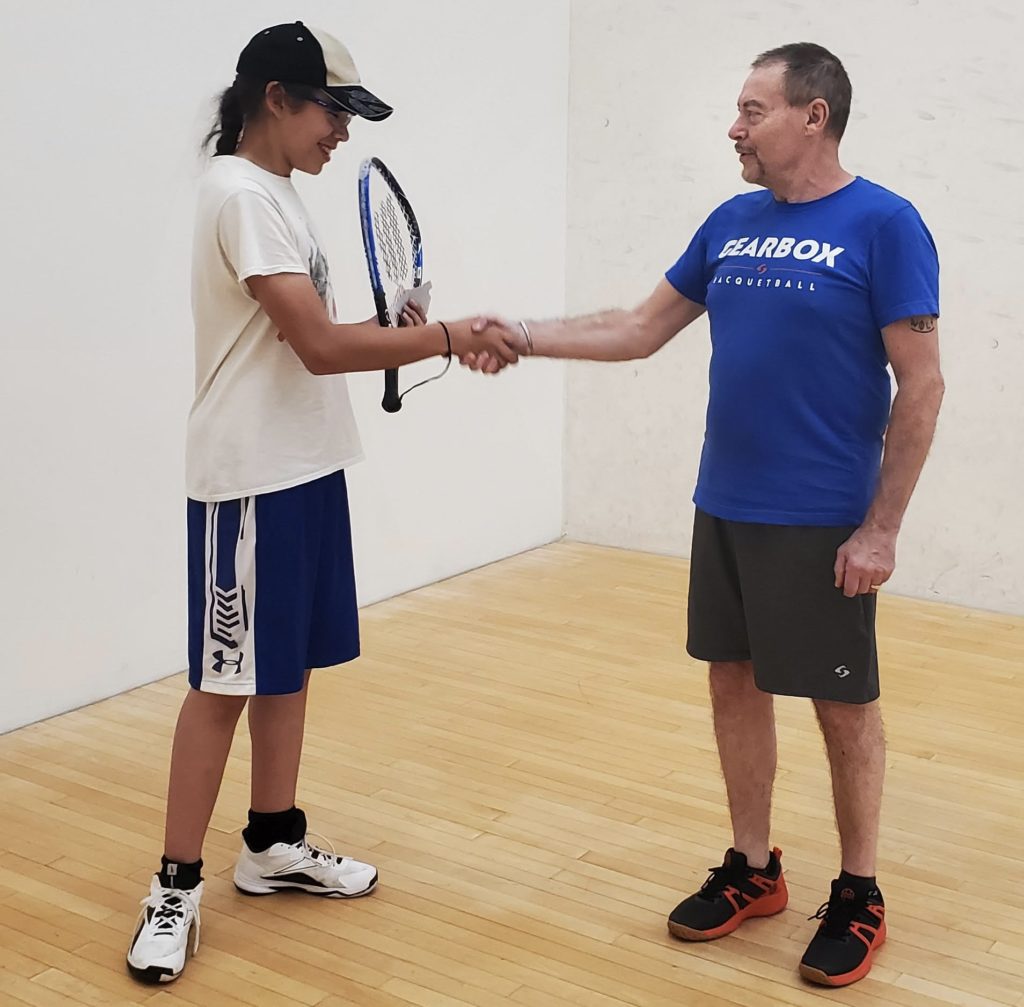 One male youth holding a racquetball racquet and a Racquetball Canada Badge Program badge shaking hands with his coach. 