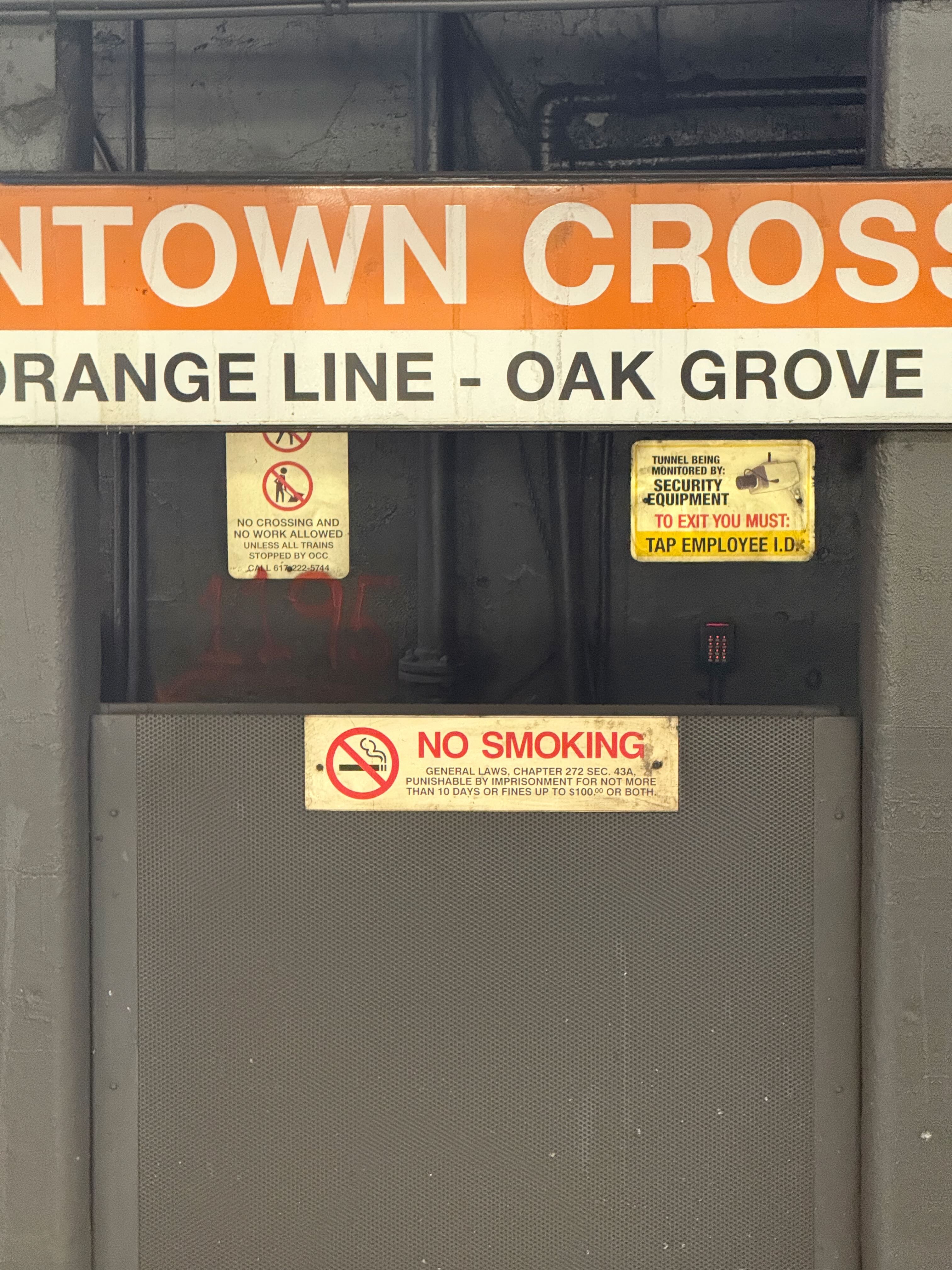 3 signs are below an MBTA Orange Line Downtown Crossing station sign. They include a sign saying “no crossing and no work allow unless all trains stopped by occ”, a sign to the right says “tunnel being monitored by security equipment, to exit you must tap employee I.D.” and the final one says “no smoking”