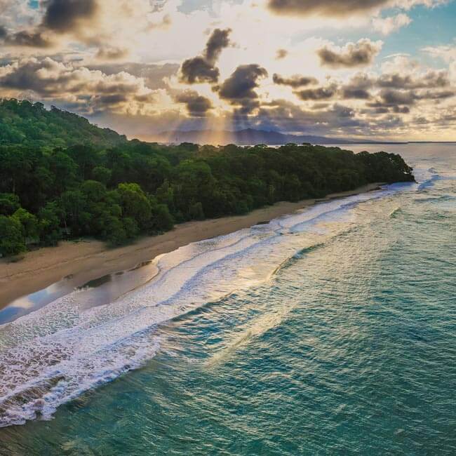 Aerial view of ocean waves gently rolling onto a sandy beach lined with dense green trees under a dramatic sky with sun rays streaming through clouds.