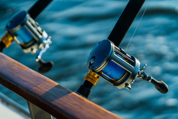 Close-up of two fishing rods with shiny reels mounted on a boat, angled over the side with blue water in the background.