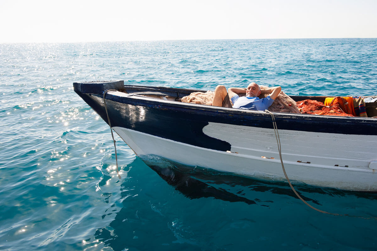 Man reclining on fishing nets inside a small boat on clear turquoise ocean waters.