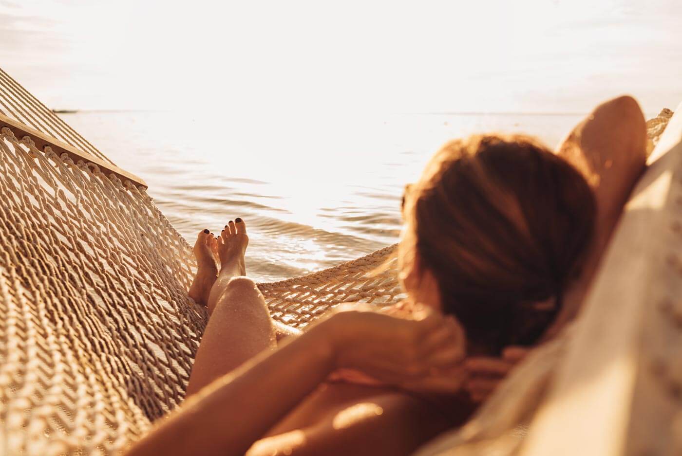 A person relaxes on a woven hammock by the water, basking in warm sunlight with an ocean or lake view in the background. Their legs are stretched out, and the scene has a peaceful, golden glow.