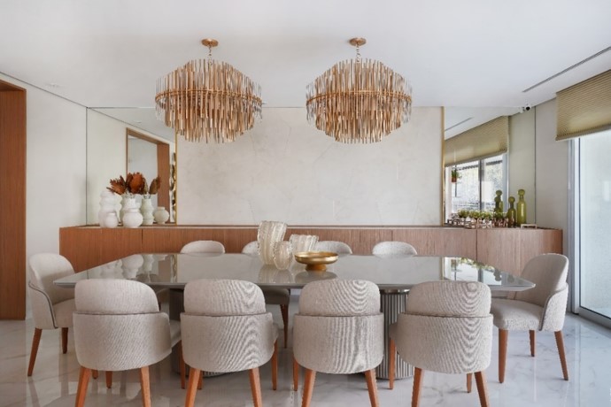Acima da mesa de jantar, o par de lustres que combinam o vidro com o dourado ornamenta a mesa com 10 lugares. Para complementar a peça ao fundo, os espelhos laterais escolhidos pela arquiteta Rosangela Pena contribuem para a amplitude da sala | Foto: Sidney Doll